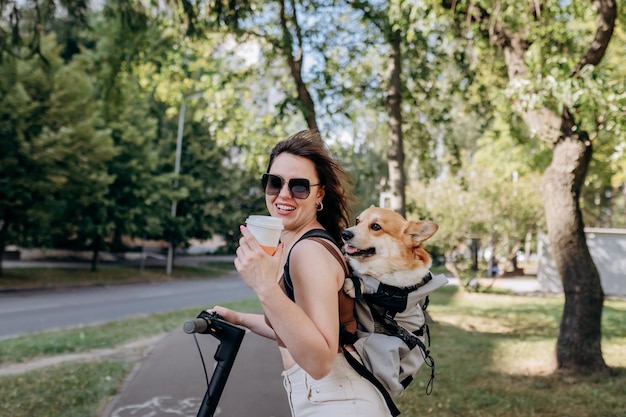 Una feliz y sonriente mujer viajera está de pie con electro scooter y bebiendo café en el parque de la ciudad con el perro Welsh Corgi Pembroke en una mochila especial