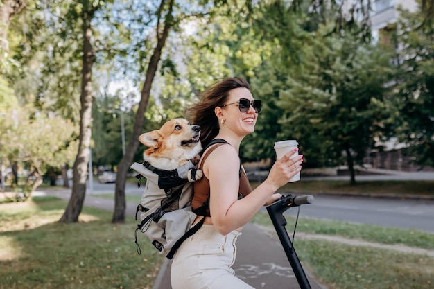 Una feliz y sonriente mujer viajera está de pie con electro scooter y bebiendo café en el parque de la ciudad con el perro Welsh Corgi Pembroke en una mochila especial