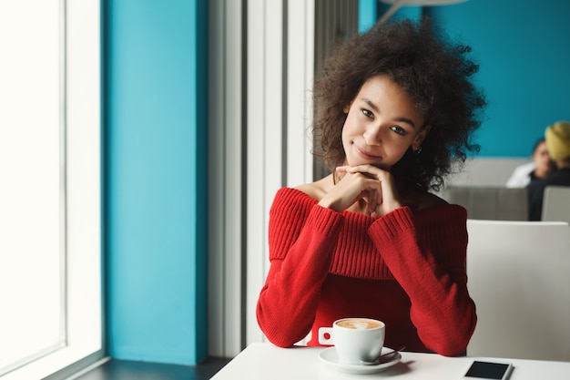 Feliz sonriente niña afroamericana en la mesa de café. Joven soñadora disfrutando de un café caliente en un ambiente agradable. Concepto de ocio y estilo de vida urbano.