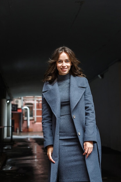 Feliz sonriente niña bonita joven en abrigo gris y cuello alto posa al aire libre. Sesión de fotos de retrato de estilo callejero de mujer atractiva, sesión de fotos urbana de hermosa modelo elegante