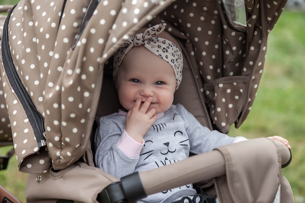 Feliz sonriente niña emocional de ojos azules de ocho meses sentarse en el cochecito de paseo y esperar a mamá. Bebé pequeño y lindo con diadema en la cabeza sentado en cochecito. Concepto de niñez y crianza adecuadas