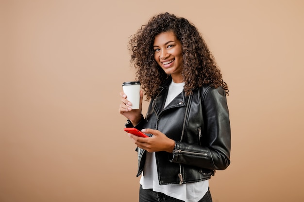 Feliz sonriente niña negra con teléfono y café para ir taza aislado sobre marrón