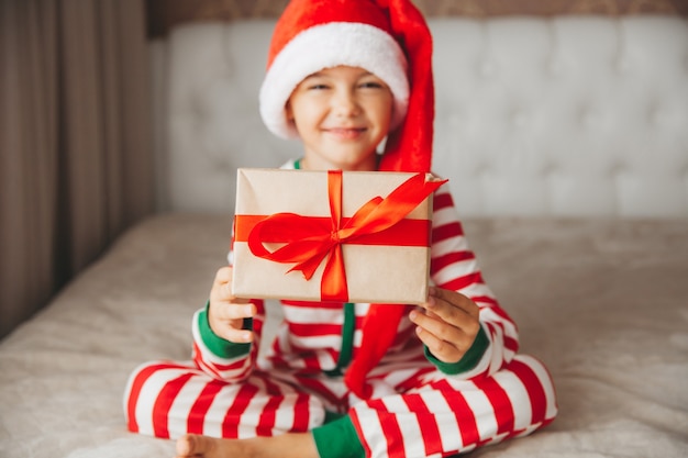 Feliz sonriente niño alegre con sombrero de Santa Claus, sosteniendo un regalo de Navidad en sus manos, sentado en la cama.