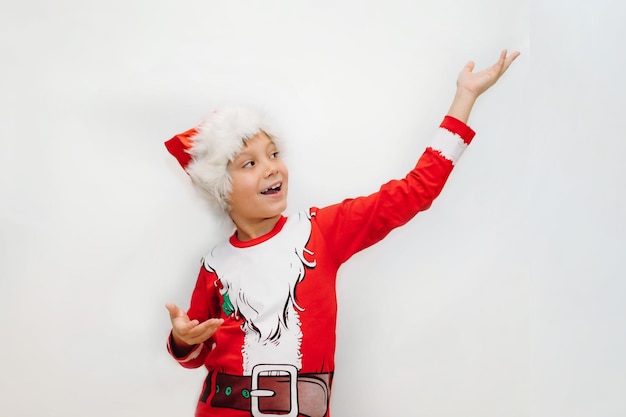 Feliz sonriente niño caucásico con camiseta de santa y sombrero apuntando al espacio en blanco sobre fondo blanco para texto publicitario