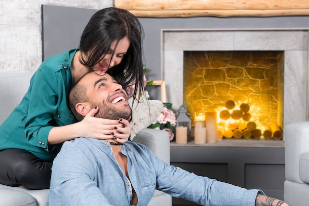 Foto feliz sonriente pareja internacional de hombre con barba y su morena esposa embarazada sentada en el sofá y abrazándolo en la sala de estar con chimenea