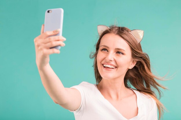 Feliz sonriente rubia adolescente hace selfie vía smartphone sobre fondo azul.