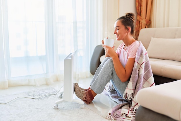 Feliz sonriente satisfecha joven alegre con calcetines a cuadros y tejidos bebiendo té caliente y calentándose cerca del calentador eléctrico en casa