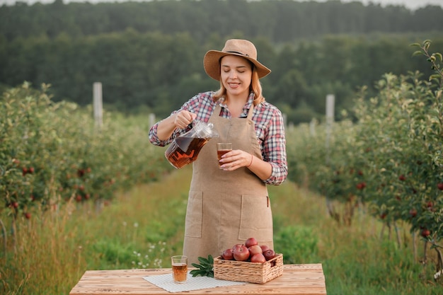 Feliz y sonriente trabajadora agricultora vertiendo sabroso jugo de manzana en vidrio de pie en el jardín de la huerta durante la cosecha de otoño Tiempo de cosecha