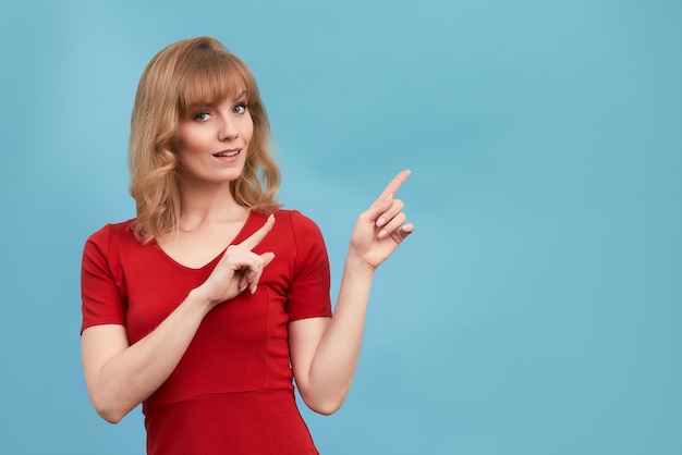 Feliz sorprendida joven rubia sonriendo ampliamente a la cámara señalando con el dedo mostrando algo interesante y emocionante en la pared del estudio con espacio de copia para su texto o contenido publicitario