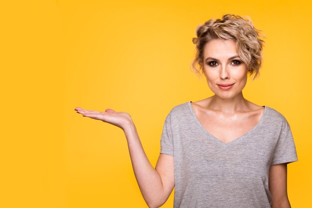 Feliz sorprendida mujer joven rubia sonriendo ampliamente a la cámara, señalando con el dedo hacia afuera, mostrando algo interesante y emocionante sobre fondo amarillo de estudio con espacio de copia para su texto