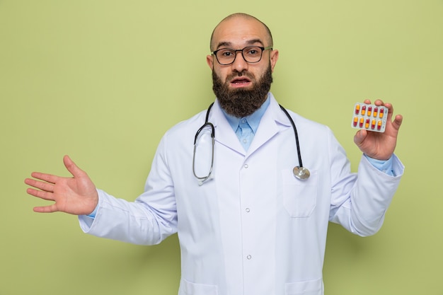 Feliz y sorprendido médico hombre barbudo en bata blanca con estetoscopio alrededor del cuello con gafas sosteniendo blister con pastillas sonriendo alegremente con el brazo levantado