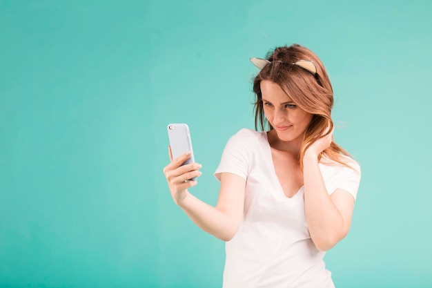 Foto feliz sorridente adolescente loira posando com telefone faz selfie via smartphone em fundo azul