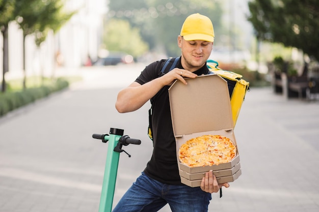 Foto feliz sorridente entregador de boné amarelo e bolsa com isolamento térmico oferecendo pizza em caixa de papelão