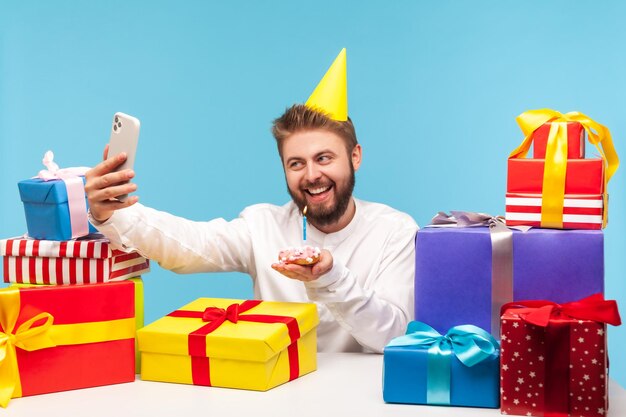 Feliz sorridente homem blogueiro no chapéu de cone comemorando seu aniversário com assinantes soprando vela no bolo sentado no local de trabalho com muitas caixas de presente Tiro de estúdio interior isolado em fundo azul