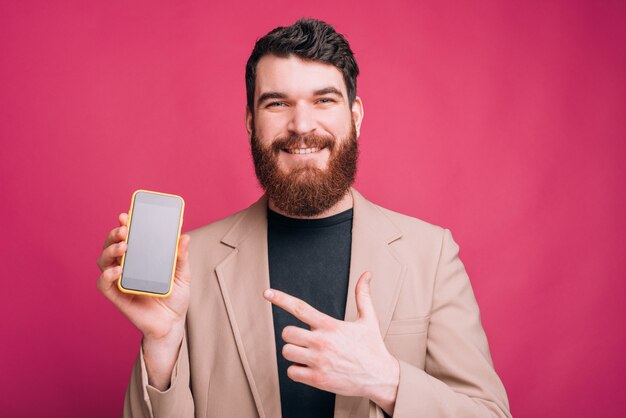 Feliz sorridente homem de terno apontando para smartphone sobre fundo rosa