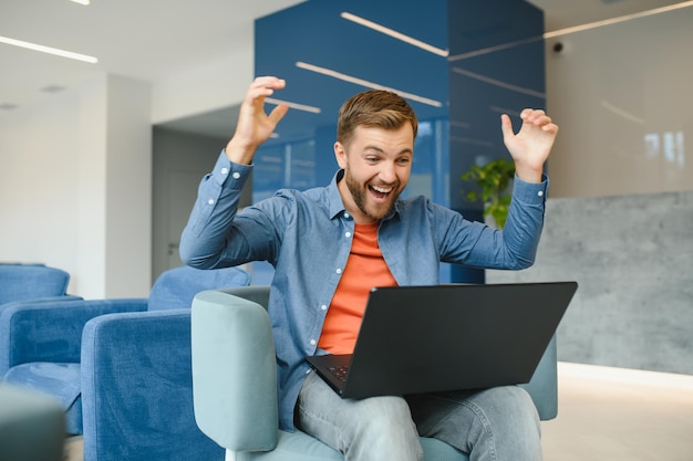 Foto feliz sorridente homem de trabalho on-line remoto em roupa casual com laptop em alegre gesto vencedor bem-sucedido sentado em um escritório de coworking em uma mesa de trabalho