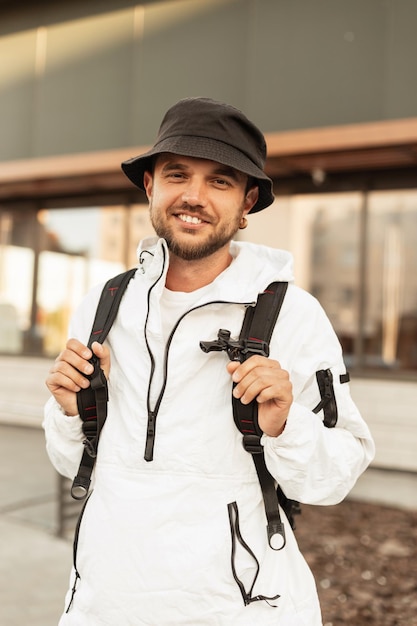 Feliz sorridente jovem hipster com chapéu em roupas urbanas de moda com mochila anda e viaja na rua