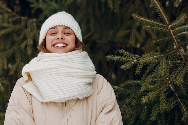 Feliz sorridente jovem retrato de mulher vestida casaco cachecol chapéu e luvas desfruta do tempo de inverno no parque de inverno com pinheiros