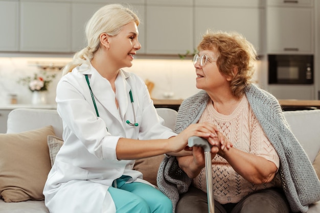 Feliz sorridente médica consolando mulher idosa segurando bastão de caminhada