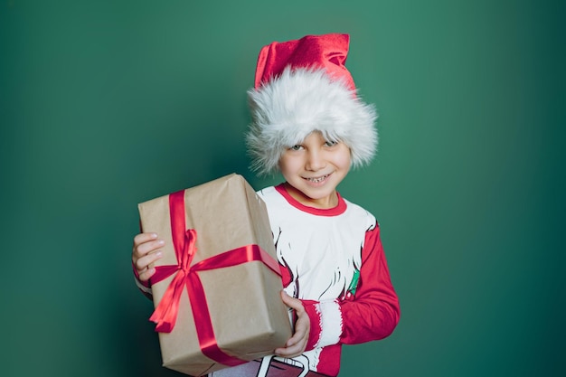 Feliz sorridente menino caucasiano em camiseta de papai noel e chapéu segurando uma caixa com presente de natal