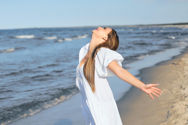 Feliz sorridente mulher bonita está na praia do oceano em um vestido de verão branco, braços abertos.
