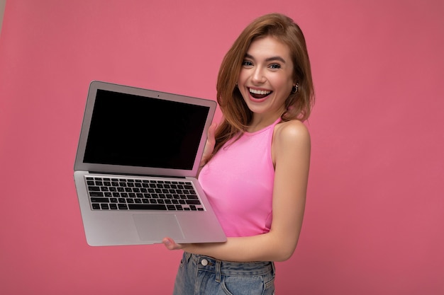 Feliz sorrindo Bela jovem loira segurando o laptop do computador vestindo a blusa rosa, olhando para a câmera isolada no fundo rosa. Simule, copie o espaço