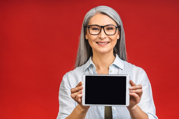 Foto feliz sorrindo feliz mulher madura asiática sênior em casual mostrando a tela do smartphone em branco enquanto olha para a câmera isolada sobre fundo vermelho usando o telefone