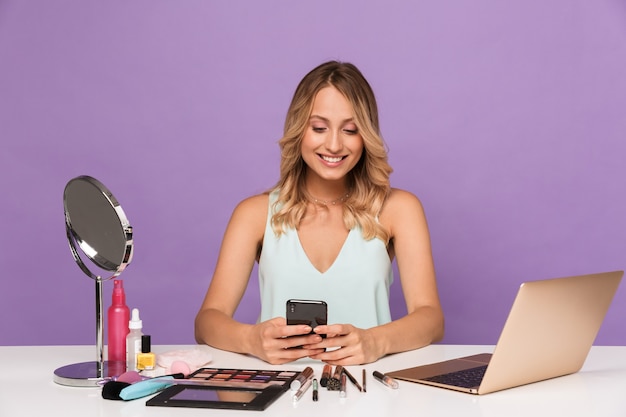 feliz sorrindo positiva jovem com computador portátil e cosméticos com espelho, usando o telefone celular.