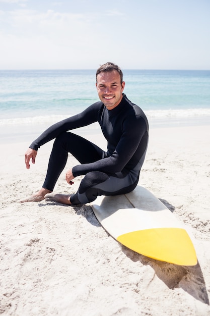 Feliz surfista en traje de neopreno sentado con tabla de surf en la playa