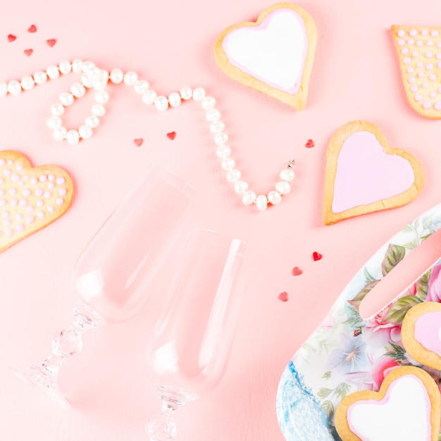Feliz tarjeta de felicitación del día de San Valentín con galletas de corazón, copas de vino y vino sobre fondo rosa pastel.