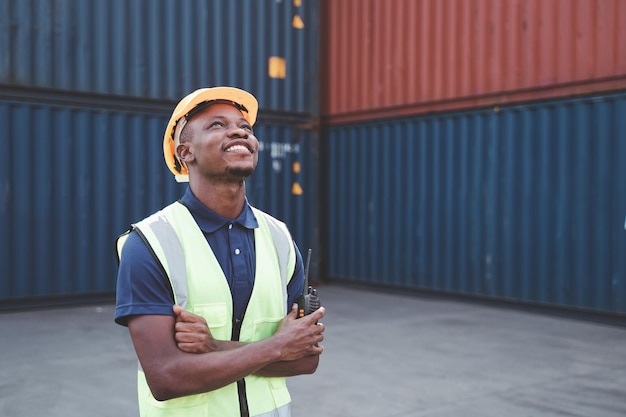 Feliz trabajador africano enviando mensajes de texto de pie en el lugar de trabajo del contenedor y cruzando los brazos con sentimiento de felicidad