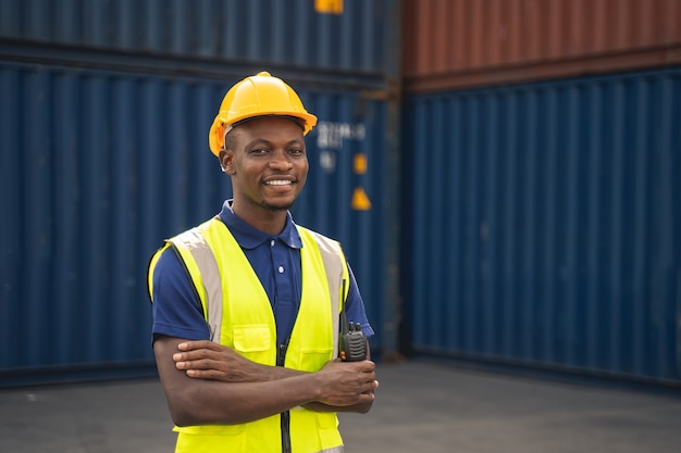 Feliz trabajador africano smailing, de pie en el lugar de trabajo del contenedor y de brazos cruzados con sentimiento de felicidad