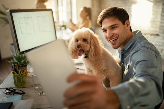 Feliz trabajador independiente divirtiéndose mientras usa el panel táctil y se toma selfie con su perro en la oficina