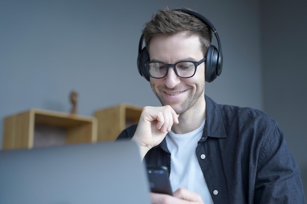 Feliz trabajador de oficina en casa con auriculares y gafas sonriendo sinceramente al mirar la pantalla del teléfono