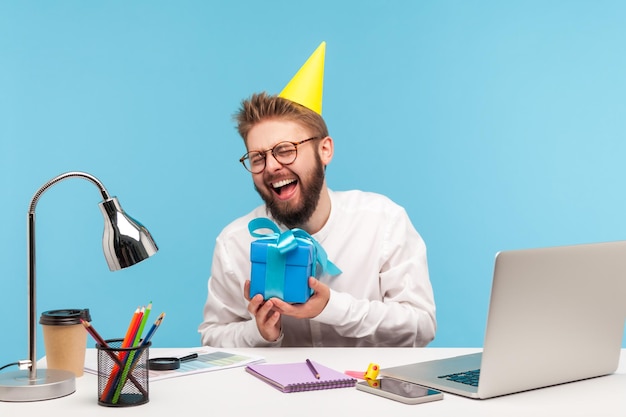 Feliz trabajador de oficina positivo con barba en camisa blanca y sombrero de cono sosteniendo una caja de regalo y riéndose sentado en el lugar de trabajo con una computadora portátil disfrutando del presente Estudio interior aislado en fondo azul