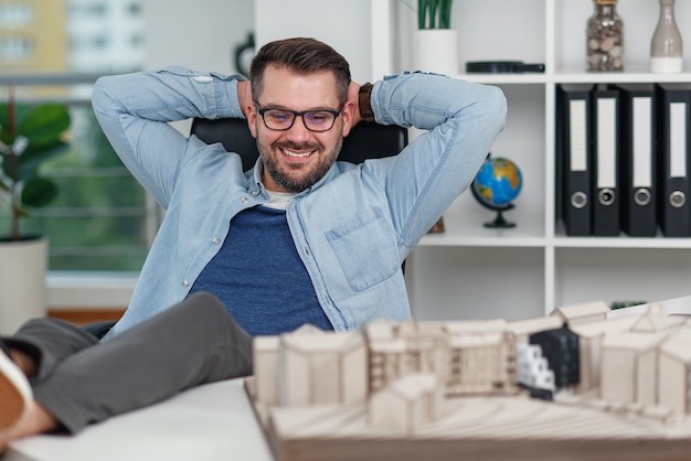 Feliz trabajador de oficina en ropa casual puso sus pies sobre la mesa del espacio de trabajo mientras soñaba con descanso o vacaciones.