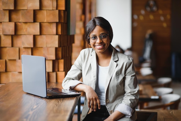 Feliz trabajadora afroamericana que usa una computadora portátil para estudiar en una computadora en una oficina o cafetería, una estudiante independiente sonriente de raza mixta que usa una aplicación de PC para comunicarse en línea viendo un seminario web