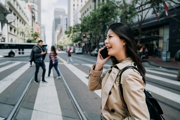 Foto feliz trabajadora asiática hablando por teléfono celular divirtiéndose charlando con amigos riendo. joven dama de oficina cruzando el tren ligero de zebra road en la ciudad de san francisco. viaje diario al trabajo en el área de negocios concurrida