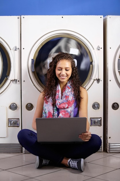 Foto feliz trabajando y una mujer con una computadora portátil en una lavandería para conexión de proyectos universitarios y multitarea educación de sonrisas y una joven estudiante leyendo en una computadora mientras lava ropa