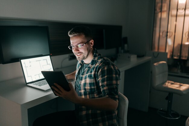 Feliz trabalhador trabalhador usando tablet e laptop enquanto está sentado no escritório tarde da noite