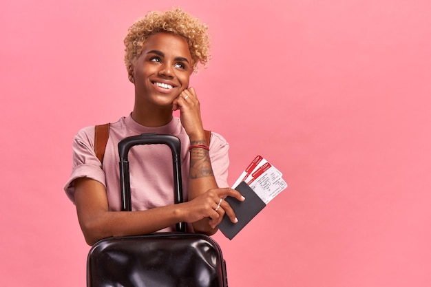 Feliz turista afro com bagagem segurando passaporte e passagens aéreas em fundo rosa