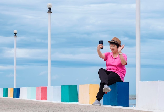 Feliz turista asiática acenando com a mão enquanto videochamada com smartphone na ponte no ponto de vista do mar