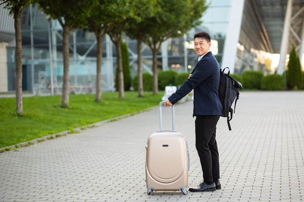 Feliz turista asiática com uma mala grande perto do aeroporto, em uma reunião de negócios chega olhando para a câmera e sorrindo