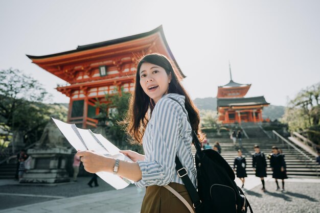 Foto feliz turista asiática feminina visitando a famosa atração turística da cidade de kyoto com mapa. jovem visitante segurando papel guiado visita templo kiyomizu em dia ensolarado. estudantes do ensino médio japonês nas costas