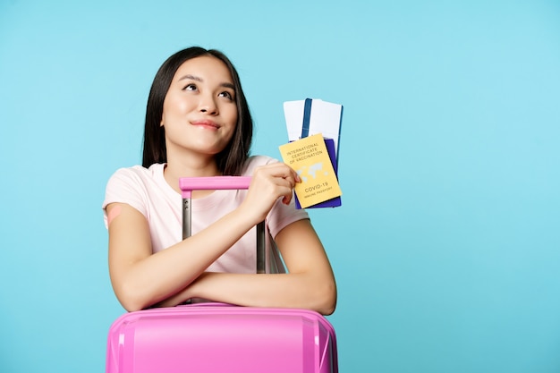 Feliz turista asiática soñando con viaje apoyarse en maleta sosteniendo la vacunación internacional covid ...