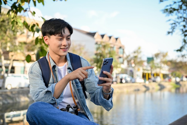 Feliz turista asiático usando seu smartphone enquanto relaxa sentado perto do pequeno canal