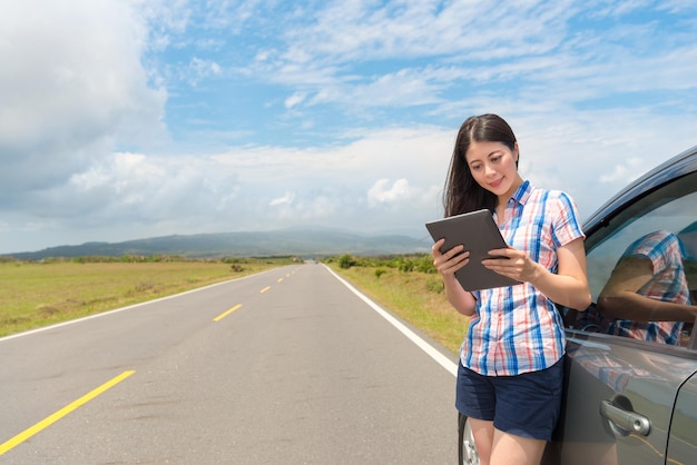feliz turista feminina elegante ao lado do carro usando o computador tablet digital móvel, encontrando o mapa procurando a rota turística na estrada de asfalto.