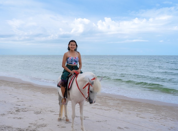 Feliz turista mujer asiática en ropa de playa a caballo blanco en la playa de arena en la playa de Huahin con mar azul y cielo nublado, relajarse de vacaciones en Tailandia