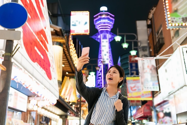 feliz turista taiwanesa que muestra la torre tsutenkaku iluminada en el fondo mientras tiene video chat con un amigo en el teléfono móvil en shinsekai new world en Osaka, Japón por la noche