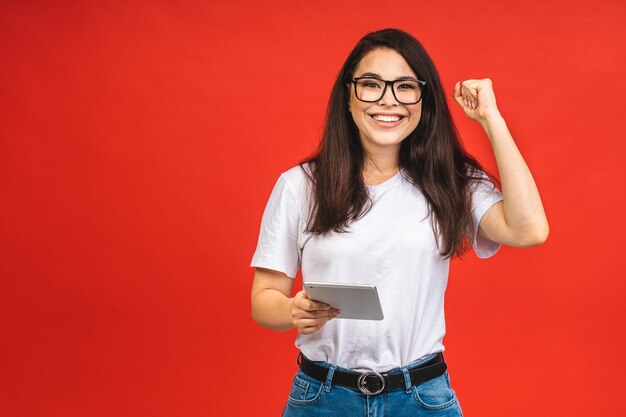 Feliz vencedor Retrato de uma jovem mulher de negócios morena sorridente usando computador tablet comemorando a vitória isolada sobre fundo vermelho